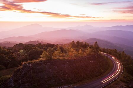 Beautiful mountain road at sunrise, curve asphalt road with light trail from headlights leading through mountain range, colorful clouds and early morning sky. Doi Inthanon, Chiang Mai, Thailand. Long exposure.の写真素材