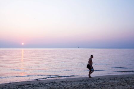 Portrait of unidentified bare-chested men with tote bag walking barefoot on a beach at sunset. Blurred motion. Long exposure. Nomad concept.の写真素材