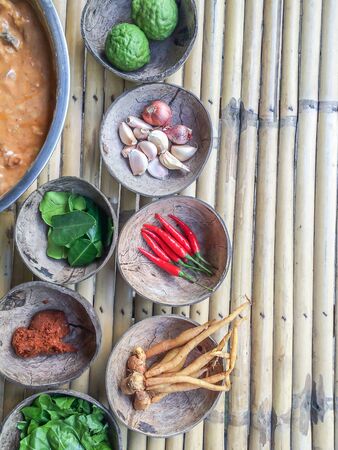 Authentic Thai red curry past recipe with ingredients in coconut shells on bamboo table, red hot chili peppers, bergamot fruits and leaves, fingerroot, ginger, lemongrass, garlic and herbs. Food culture. Top view.の写真素材