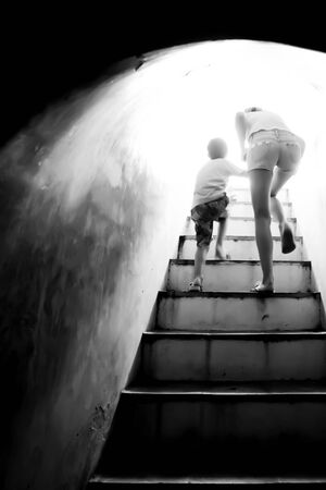 Motion blur, out of focus. Mother holding hand her son walking up the stairs to bright sun light at the end of dark tunnel. Monochrome. Freedom concept.の写真素材