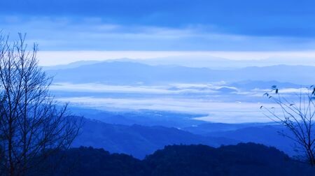 Fantastic blue clouds and sunrise sky over blue mountain range, blur neon light dots in the misty valley, bare branches of wild trees on foregrounds, remote location at Thailand-Myanmar border.の写真素材
