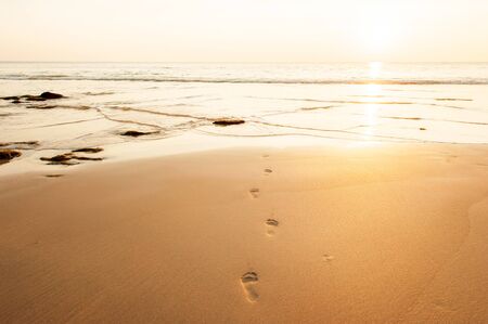Human footprints on the golden sand beach at dusk, vanished traces of feet in the ripple waves, the sun setting on the sea backgrounds, summer scene in Lanta Island, Thailand.の写真素材