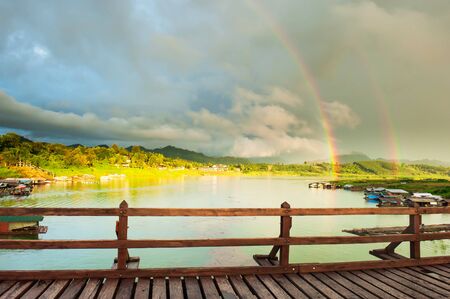 Colorful double rainbows over the lake and houseboat community on rainy day, view from old wooden bridge over the lake. Mon Thai community of Sangkhla Buri, Thailand.の写真素材