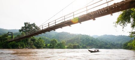 A local wooden boat passing the suspension wooden bridge at dusk, remote location in deep jungle near Thailand-Myanmar border. Motion blur.の写真素材