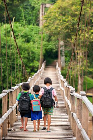 Asian primary school girls in casual clothes walking cross a simple suspension footbridge at dusk, rural scene near Thailand and Myanmar border. Education concept.の写真素材