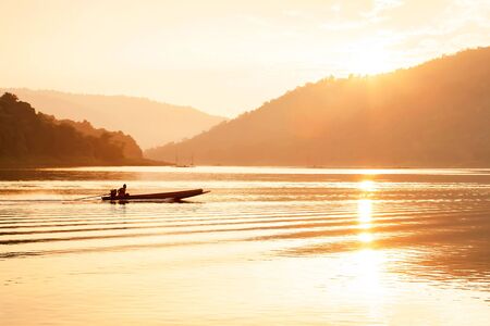 Male asian fisherman steering wooden fishing boat on the lake at dusk, the sun setting behind...