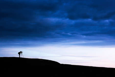 Side view shot of male photographer is taking images with dslr camera and tripod on the mountain peak under dramatic dark storm sky. Exploration concept.のeditorial素材