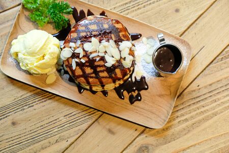 Top view of banana pancakes with macadamia nut and chocolate syrup topping on stack of pancakes and vanilla ice cream on wooden plate on wooden table. Focus on macadamia nut.の写真素材