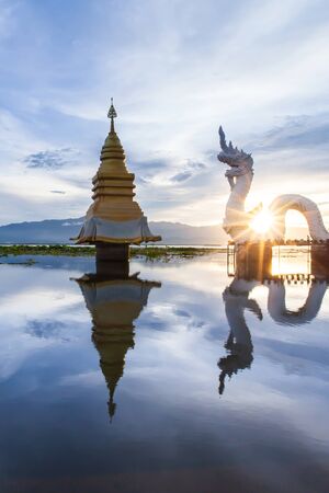 Naga statue and golden stupa on the lake at dusk, glowing sun setting through the Naga statue. Phayao Lake, Thailand.の写真素材