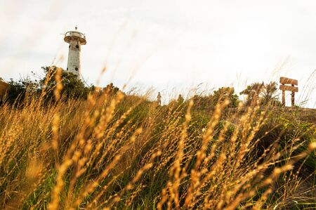 Scenery of golden grassland and ancient lighthouse on the hill at sunset, tourist family relaxing by the sunset view in front of the lighthouse, a little boy is sitting on his fatherâs shoulders. Krabi, Thailand.の写真素材