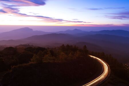 Beautiful mountain road at dawn, curvy mountain road with light trails against sunrise sky in the background. Long exposure.の写真素材