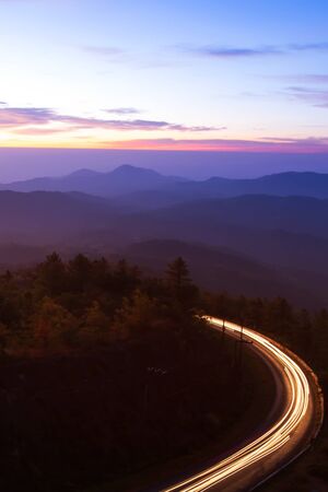 Beautiful mountain road at dawn, curvy mountain road with light trails against sunrise sky in the background. Long exposure.の写真素材