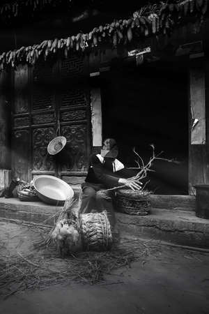 A Chinese old man making corn chair at his yard, corn cob hanging on wooden lintel of ancient house. Chengzi Village, Yunnan, China. Focus on old man.の写真素材