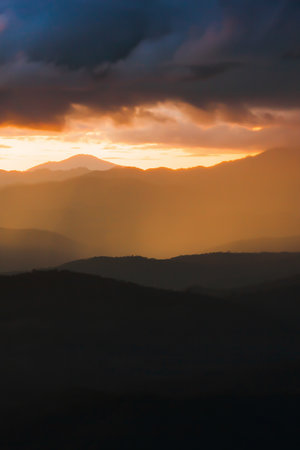 Scenery mountain range at sunset, dramatic rainstorm hiding the setting sun in the background, magical spotlight shines through dark clouds onto mountains. Focus on mountains.の写真素材
