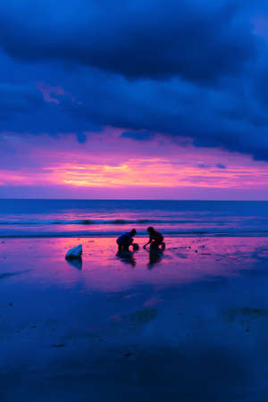 An Asian family catching fish with fishing net at a sunset beach. Dramatic dark storm clouds in the...