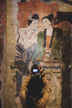 A tourist girl taking pictures of an ancient mural painting of man whispering to woman by digital camera. Wat Phumin, a famous buddhist temple in Nan, Thailand. The temple is open to the Public. Focus on mural.のeditorial素材