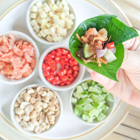Close-up hand holding royal leaf wrap appetizer or Miang Kham, authentic Thai snacks, dried salted prawn, ginger, chili, lemon, roasted peanuts, ingredients flat lay in the background. Thai food. Top view.の写真素材
