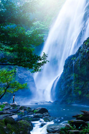 An Asian barechested man stands on the rock and looks at the great waterfall in rain season. Khlong Lan National Park, Thailand. Vacations, relaxation concepts. Long exposure.の写真素材