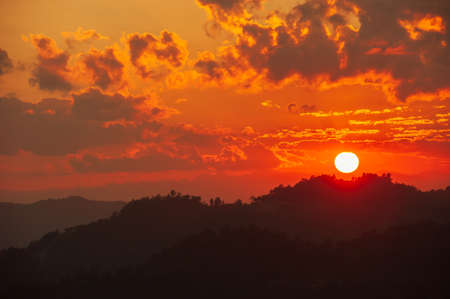 The glowing sun setting over mountains range near Thailand-Myanmar border. Dramatic clouds in the sunset sky. Silhouette.の写真素材
