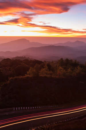 The scenery of mountain road at dawn, curve mountain road with lights trails, dramatic clouds in the sunrise sky in the background. Transportation, travel concepts. Long exposure.の写真素材