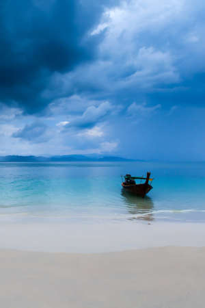 A long-tail boat moored while waiting for tourists on an island beach during monsoon, a dark rainstorm covers the Andaman Sea in the background. South Thailand. Environment, tourism concepts.の写真素材