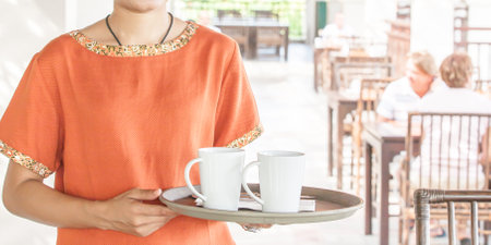 Close-up of an Asian girl waitress standing in an open-air restaurant or cafe holding a tray to serve cups of coffee to customers sitting on chairs blurred in the background.の写真素材