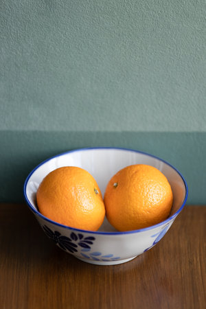 Two oranges fruit on a white ceramic bowl on the brown teak table, dark green wall in the background.の写真素材