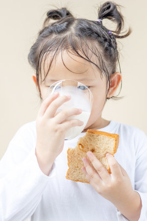 An Asian preschool girl is wearing a white long-sleeved T-shirt and drinking fresh milk from a glass with a slice of bread isolated in the background.の写真素材