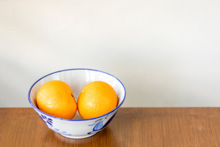 Two oranges on a white ceramic bowl on a brown table, cream wall in the background.の写真素材