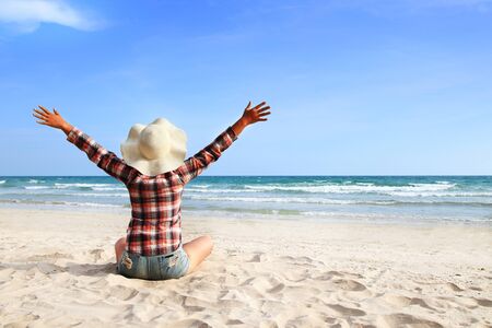 A woman wearing a plaid shirt raises her hands showing a refreshing on sky at a beautiful beachの写真素材