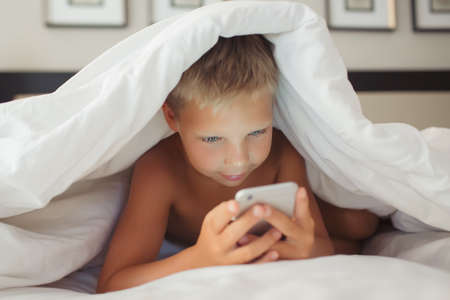 A beautiful white boy lies in bed under a blanket and holds a gadget in his hands.の写真素材