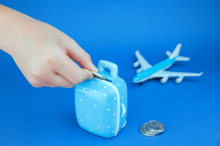 Hand putting coin into suitcase shaped money saving box and toy airplane, blue on blue backgroundの写真素材
