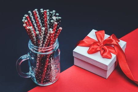 white gift box with red ribbon on black and red background with black red and white dotted paper straws in glass jar. Celebration and gift concept beyond plastic party with copy spaceの写真素材