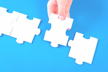 Large white blank Jigsaw puzzle matching pieces. Hand holds one piece. Teamwork and solution or idea and education learning concept with copy space on trendy blue backgroundの写真素材