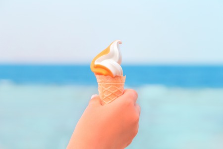 Hand of a child holding an ice cream cone with ice cream against blue sea. Summer heat fun and...