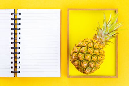 Open notepad and baby pineapple inside a wooden frame on bright warm glowing summer yellow background. Exotic concept with copy spaceの写真素材