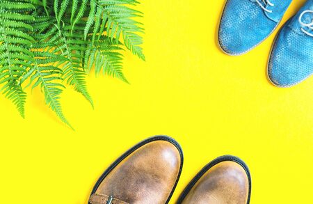 Autumn or spring trendy flat lay on bright glowing yellow. Two pairs of shoes brown male and blue female and fresh green fern leaves. copy spaceの写真素材