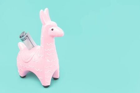 Pink zine type toy alpaca llama with dollar note on blue background close up, coin money bank. Creative and fun trendy collage of funky animal saving for travel concept with copy spaceの写真素材