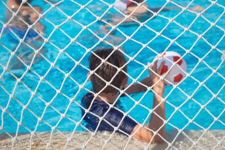 Children paly water polo with ball in swimming pool. View through goal net selective focus. Summer fun sports with copy spaceの写真素材