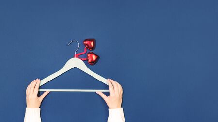 Female hands holding empty coat hanger with two red glass heart pendants. Flat lay on dark blue. 14...