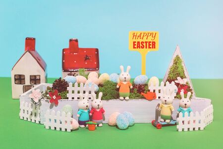 Happy Easter sign and a toy house of bunnies family in the garden and dyed eggs hunt. Greeting card concept.の写真素材