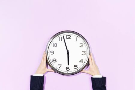 Two Hands of woman in business suit hold big plain wall clock on pastel pink background. six oclock....