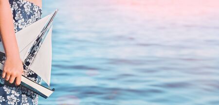 Child caucasian tanned boy in swimming trunks at the seaside beach holding toy sailing boat. Blue sunny waters. Exotic travel and destination scenic vacation web banner with copy spaceの写真素材