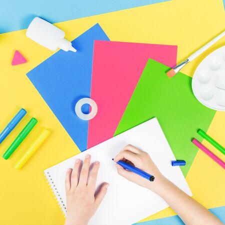 Hands drawing over color paper, scissors, pencils paint and various school stationery on blue yellow background. Flat lay with copy space for back to school or education and craft conceptの写真素材