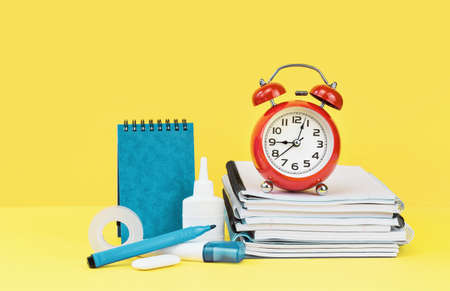 Sketchbook and note pads with office supplies and school blue stationery and red alarm clock on yellow desk. back to school or education and craft concept. Alarm Clock at 9 oclock.の写真素材