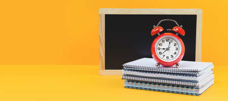 Pile of notebooks school chalk blackboard and red alarm clock on orange desk. back to school or...