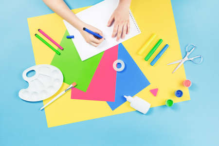 Hands drawing over color paper, scissors, pencils paint and various school stationery on blue yellow background. Flat lay with copy space for back to school or education and craft conceptの写真素材