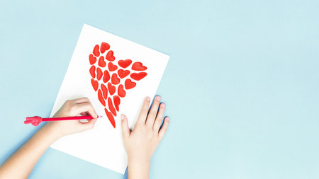Hands on desk with a red pencil and a heart shape on white sheet on blue background. Flat lay with copy space. Love letter for St Valentine's or Mother's Day. Banner for saleの写真素材