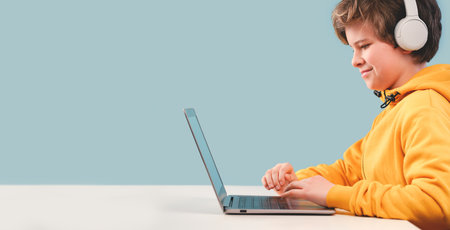 Child caucasian boy in yellow hoodie with headphones studying at home with laptop. Remote or distant...