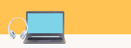 Laptop. Mockup screen and headphones on white desk and plain yellow background banner. Distant learning. working from home, online courses or support minimal concept. call center headsetの写真素材